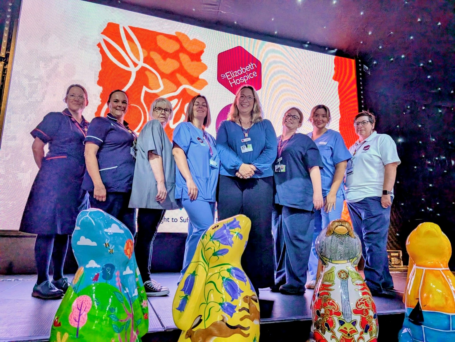 Group photo of St Elizabeth Hospice staff in uniform standing on stage with hare sculptures, in front of the hospice logo at the auction event